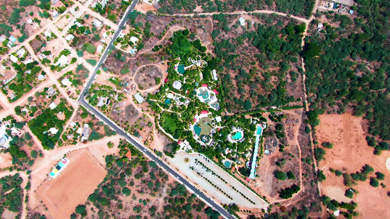 Top-down Caduano frame showing the road beside a cluster of blue pools, dense landscaping, parked vehicles, nearby homes, dry open ground, and no visible people.