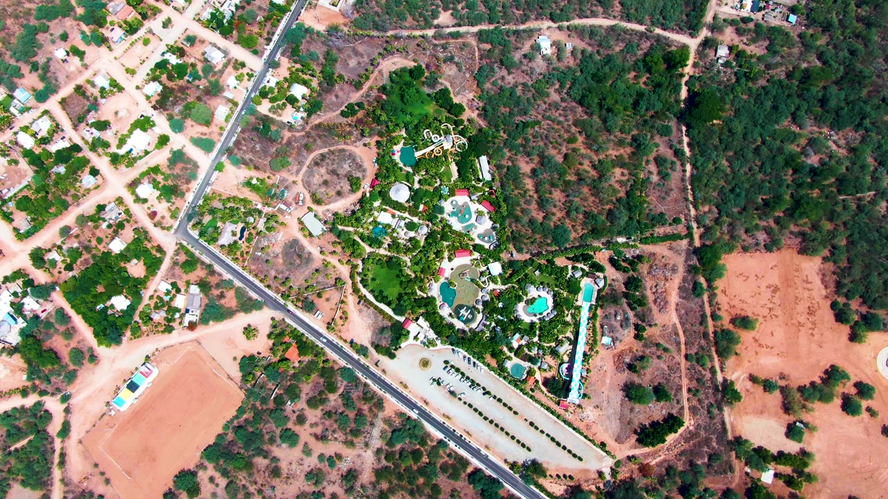 Overhead frame of a paved road dividing residential plots, resort greenery, and dry open land.
