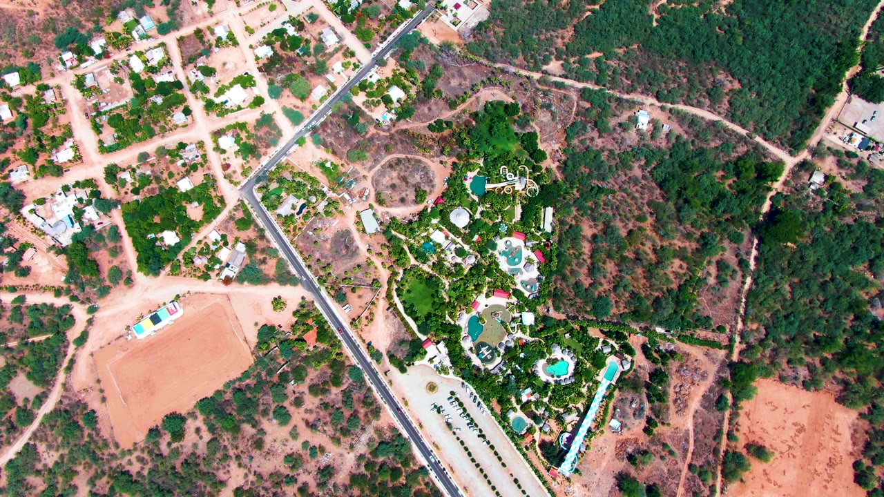 Opening overhead frame in Caduano, Baja California Sur, showing a diagonal paved road, scattered houses, blue pools, dense trees, reddish dirt fields, and no visible people.