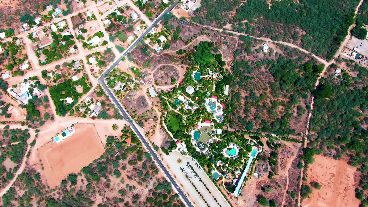 Aerial frame of a road, neighborhood lots, pools, trees, and dry open land.