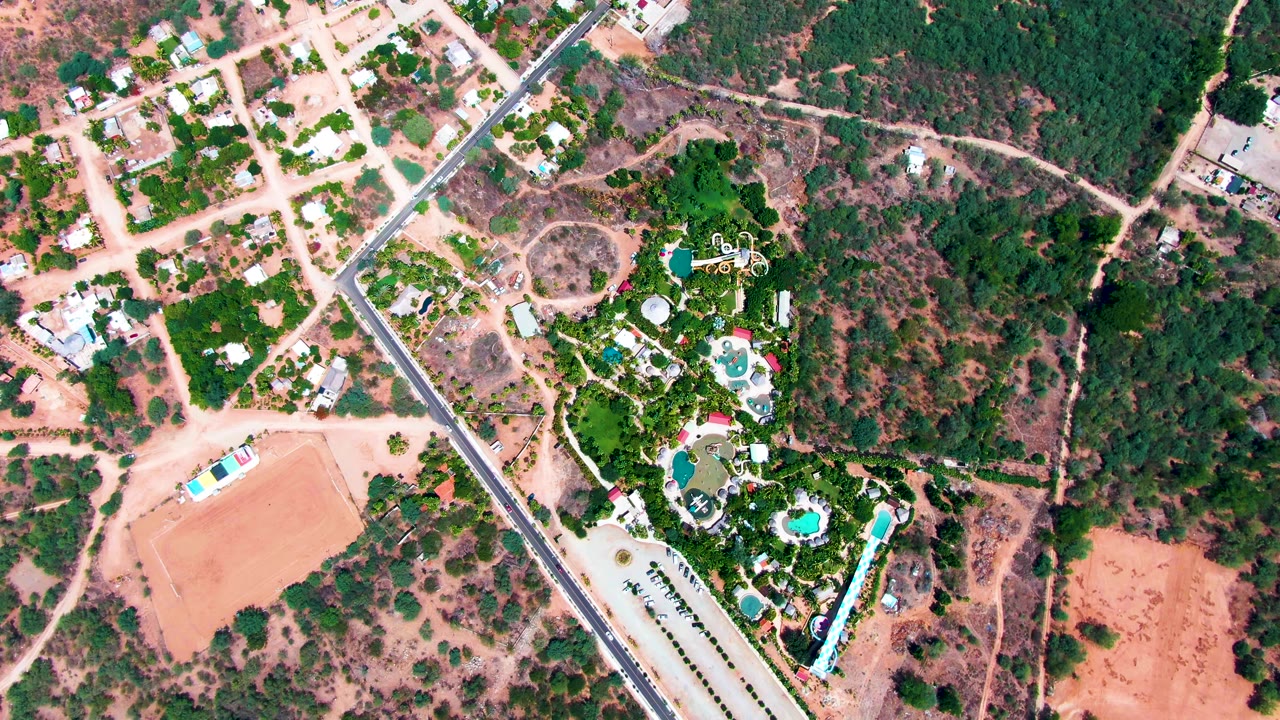 At 0:02, the Caduano overhead angle shows the same diagonal road bordered by homes, pool shapes, green landscaping, scrubby trees, dry lots, and no visible people.