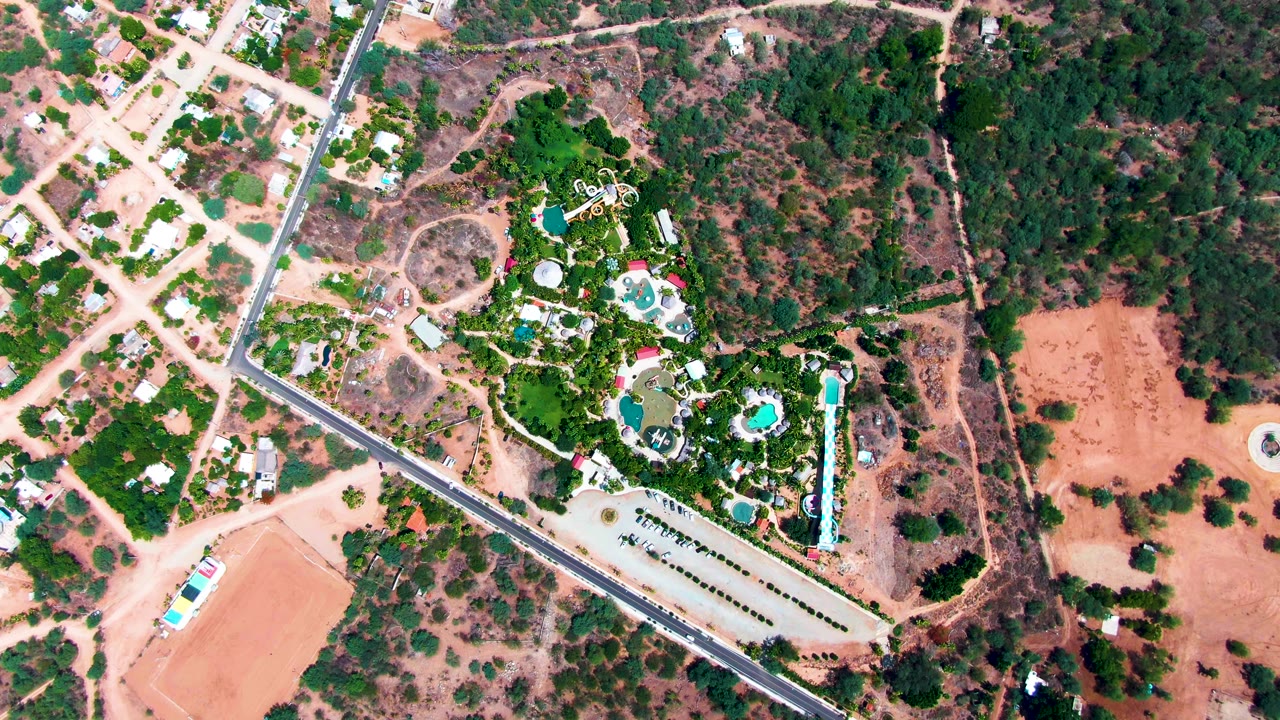 Overhead aerial frame in Caduano, Baja California Sur showing a green estate with blue pools, surrounding roads, nearby houses, reddish dirt parcels, and no visible people.
