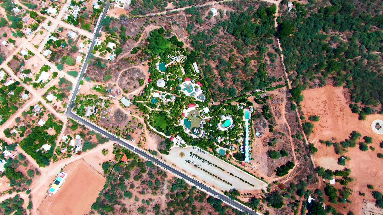 Top-down frame in Caduano, Baja California Sur showing turquoise pool shapes inside dense greenery, a straight road along the estate edge, dry lots, scattered homes, and no visible people.
