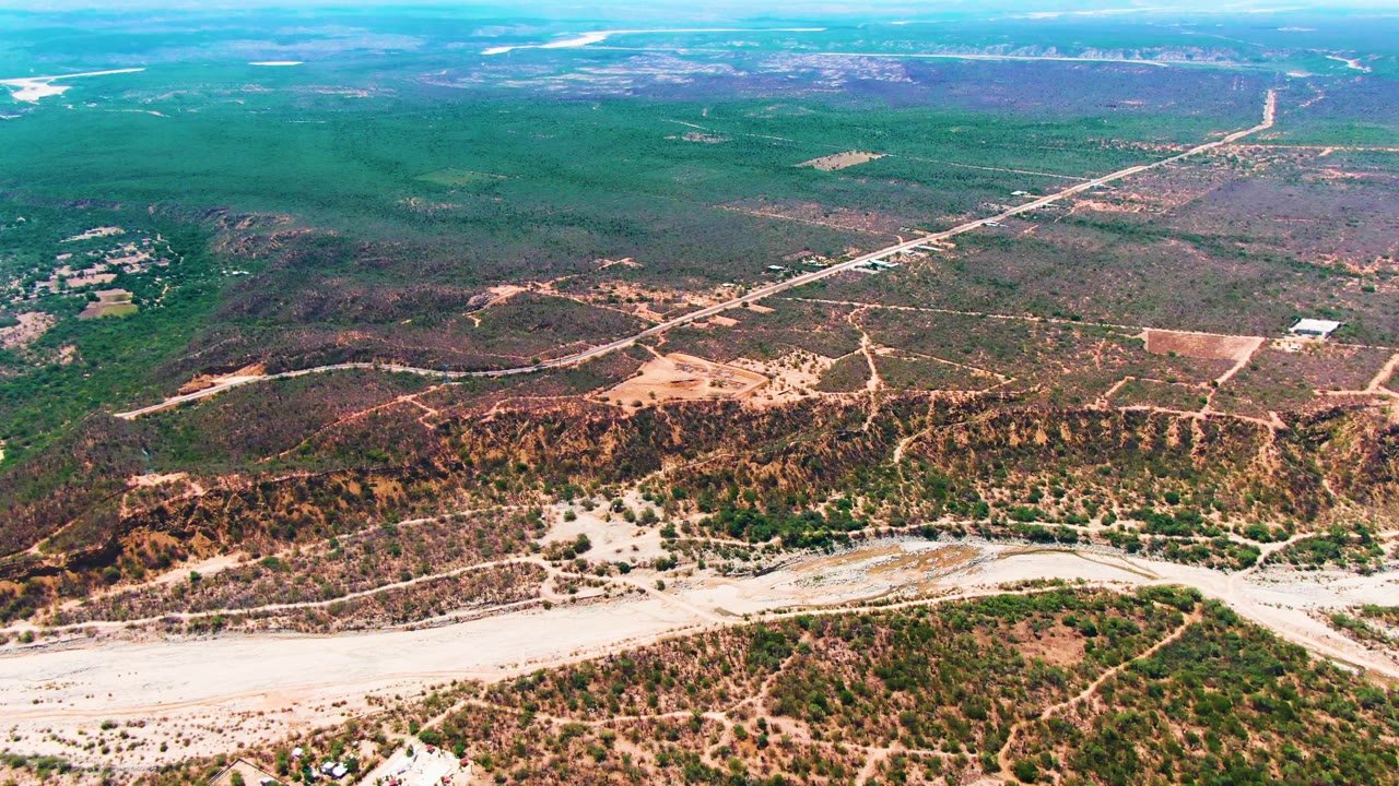 Aerial view of dry hills, riverbed, scrub vegetation, and a road in the distance.