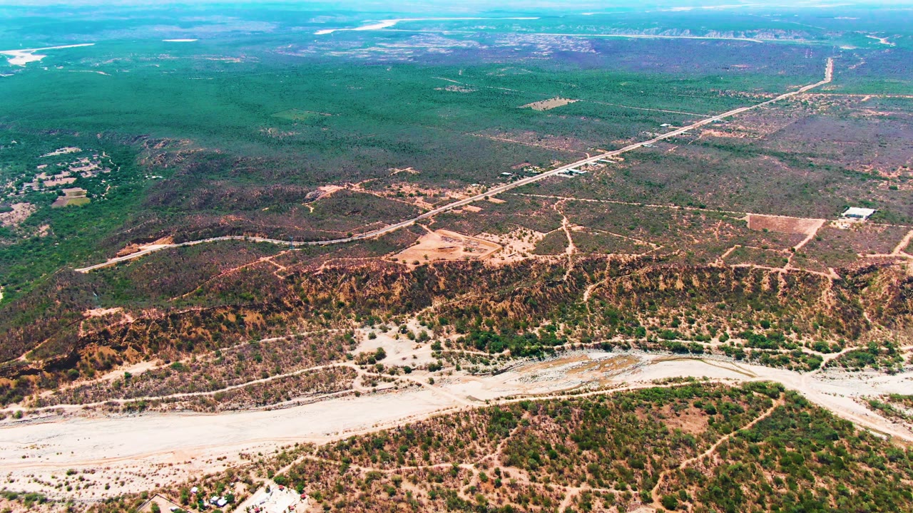 Aerial view over Caduano with the pale riverbed stretching across the lower frame, dry green-brown hills beyond it, a long road crossing the upper landscape, and no visible people.