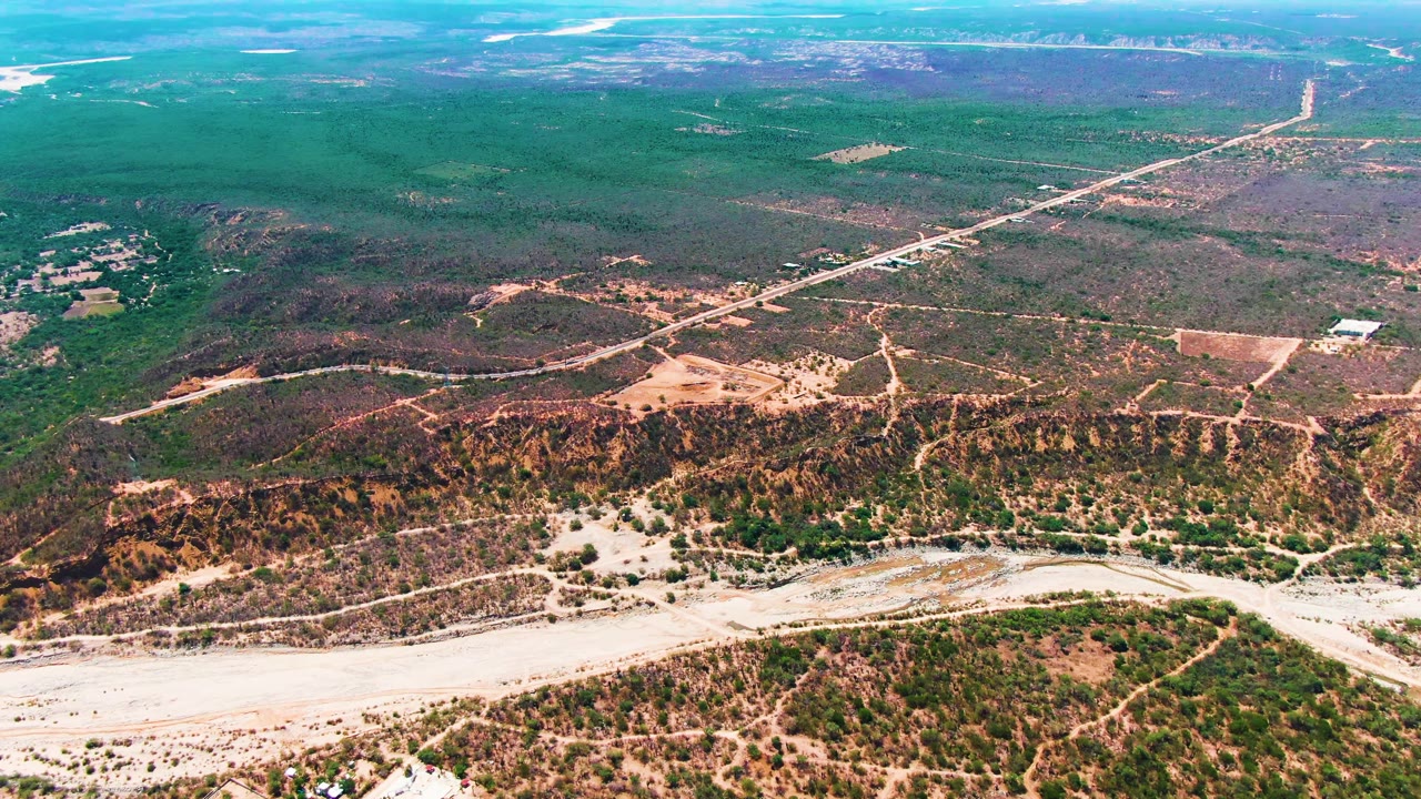 Caduano aerial frame showing a winding pale riverbed at the bottom edge, mottled scrub on low hills, a straight road cutting through the upper right, and no visible people.