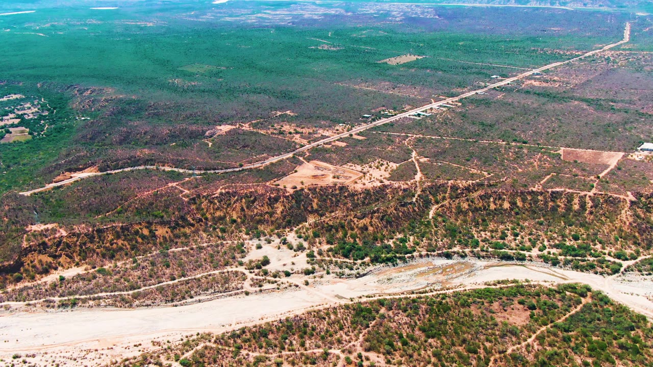 Mid-clip aerial view in Caduano with the riverbed still across the lower foreground, rough hill slopes in the midground, dirt tracks, a distant road corridor, and no visible people.