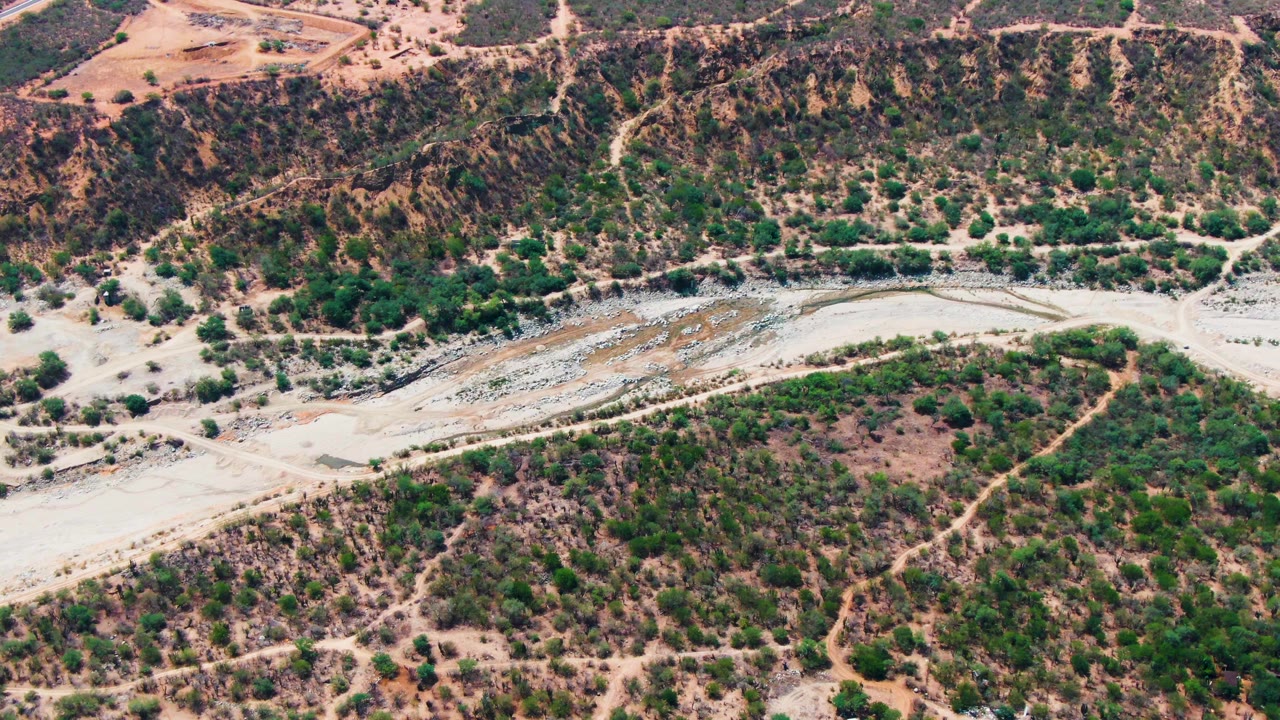 A high aerial frame over Caduano, Baja California Sur shows a pale dry riverbed curving through brown scrubland with green shrubs and no visible people.