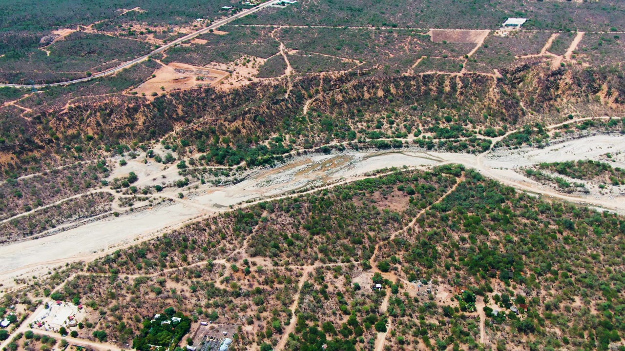 High aerial view over Caduano, Baja California Sur, showing a pale dry wash, scrub-covered hills, dirt tracks, sparse buildings at the lower edge, and no visible people.