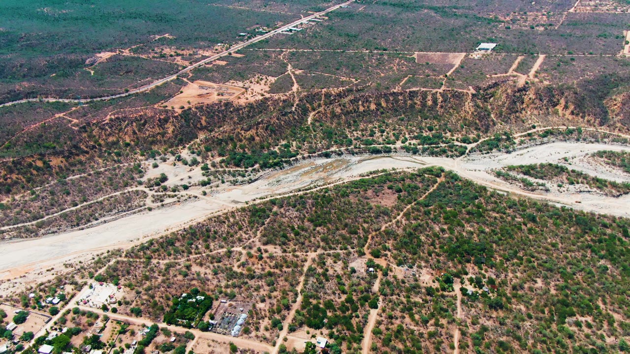 Wide drone view in Caduano, Baja California Sur, showing the pale riverbed across the scene, a straight road near the horizon, scattered structures below, and no visible people.