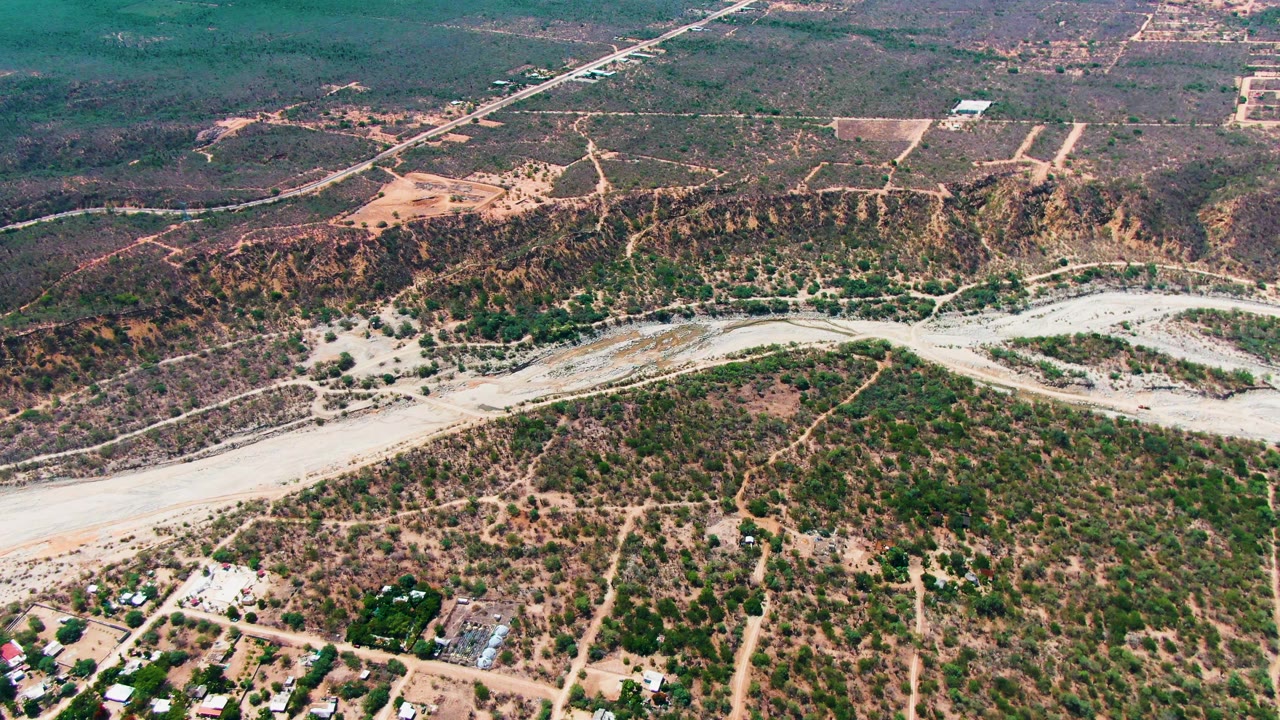 High oblique view of Caduano, Baja California Sur, with tan dirt paths through scrub, a pale wash across the midground, buildings partly visible at the lower edge, and no visible people.