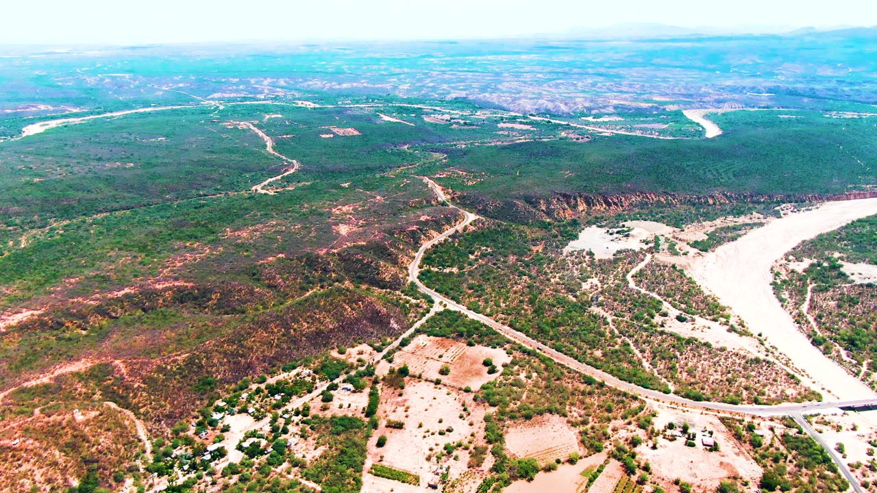 High aerial view over Caduano, Baja California Sur, showing a pale winding riverbed, branching roads, green and brown hills, distant plains, and no visible people.