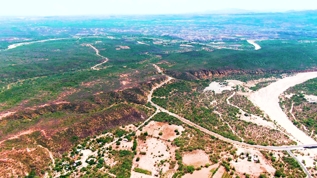 Aerial frame from Caduano with a dry-looking light river channel curving through hilly vegetation, roads cutting across the midground, distant flat land, and no visible people.