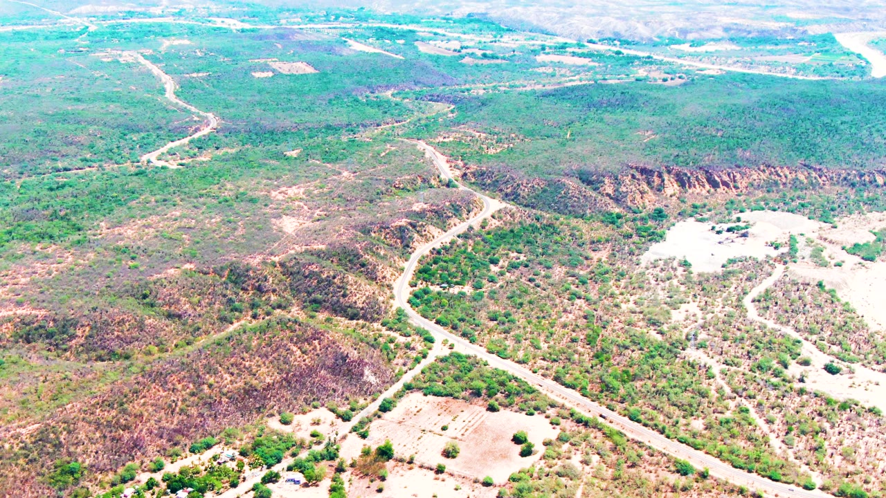 A high aerial view over Caduano, Baja California Sur shows a pale winding road crossing scrubby green and tan hills, with no visible people.