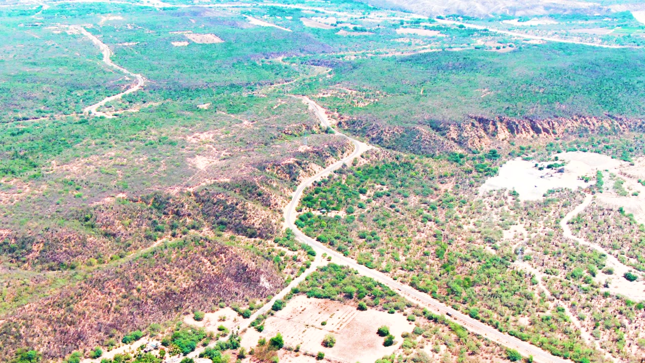 An aerial frame in Caduano, Baja California Sur holds on the curving road as it passes between rocky slopes and pale open ground, with no visible people.