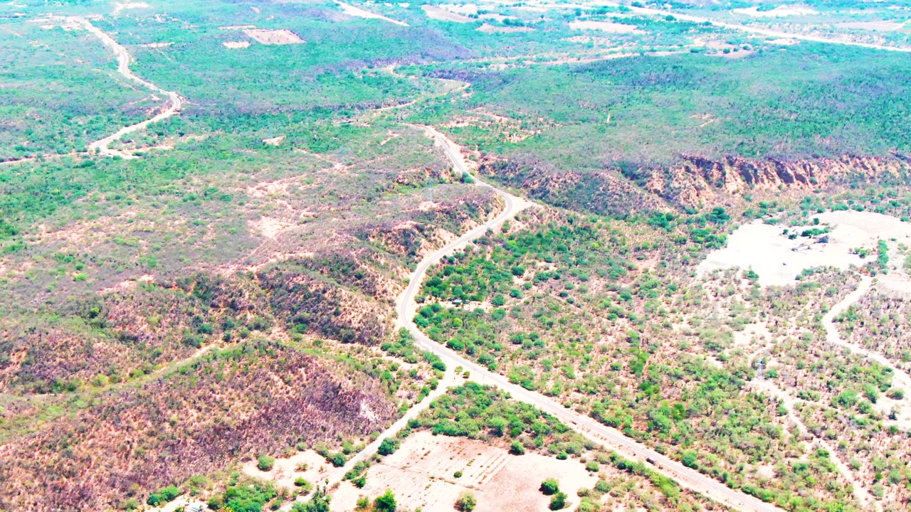 The aerial view over Caduano, Baja California Sur shows the road bending through low hills, scattered shrubs, and bright exposed soil, with no visible people.