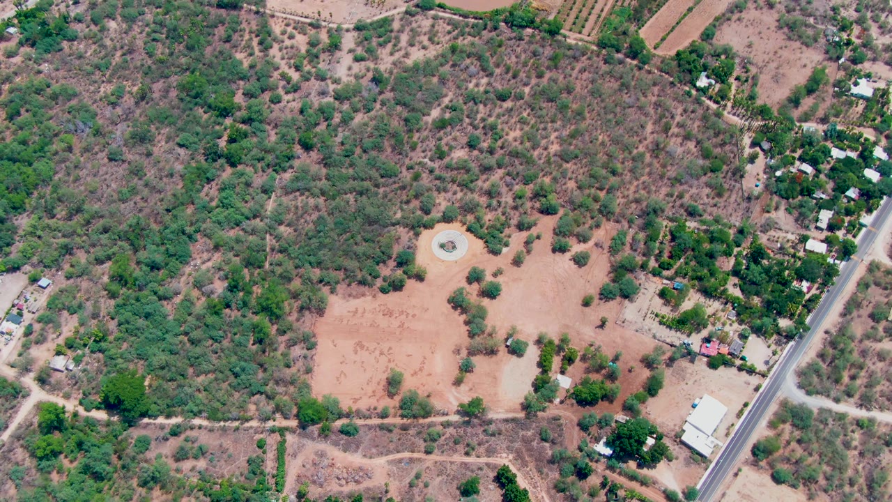 Early aerial frame over Caduano, Baja California Sur showing a tan dirt field, scattered trees, small buildings, dirt tracks, and a paved road on the right with no visible people.