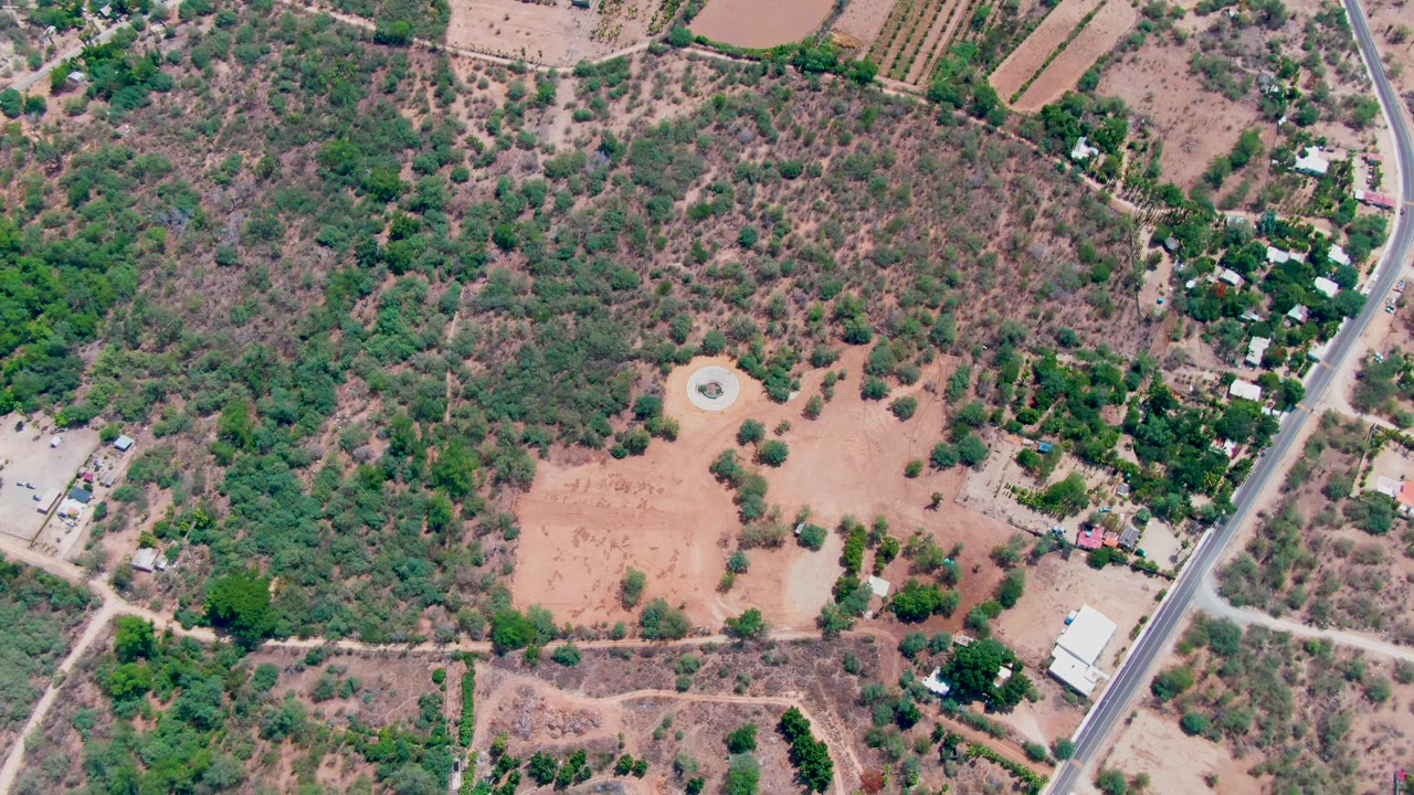 Overhead rural landscape with dry field, circular feature, trees, buildings, and road.