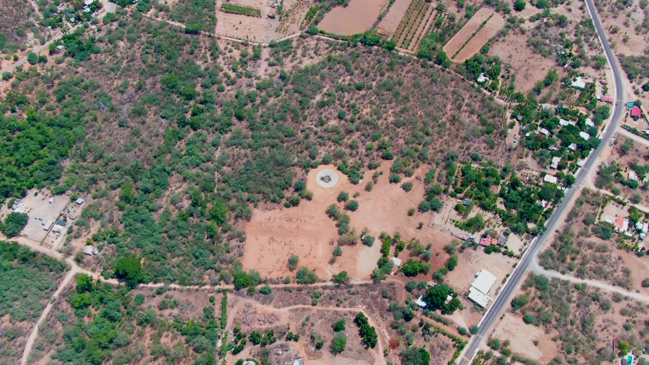 Later overhead frame in Caduano, Baja California Sur showing dry scrubland around the open field, rectangular farm plots near the top edge, roadside buildings, and no visible people.
