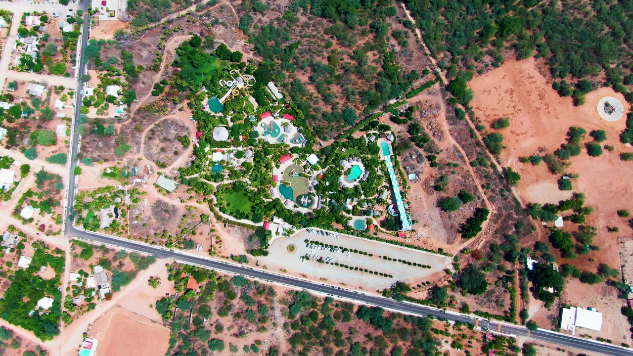 Drone frame over Caduano with the pool compound centered, the road and parking area below, scrub-covered hillside above, dry open land to the right, and no visible people.