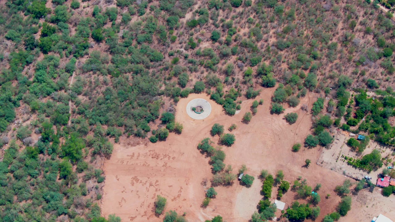 Aerial frame in Caduano, Baja California Sur with the circular structure near center, dry vegetation across the hillside, small structures at right, and no visible people.