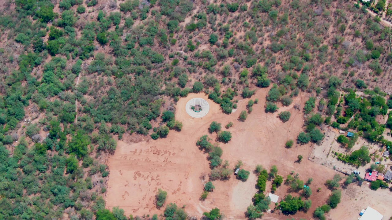 Overhead view of sparse vegetation, reddish dirt, and a circular structure near center.