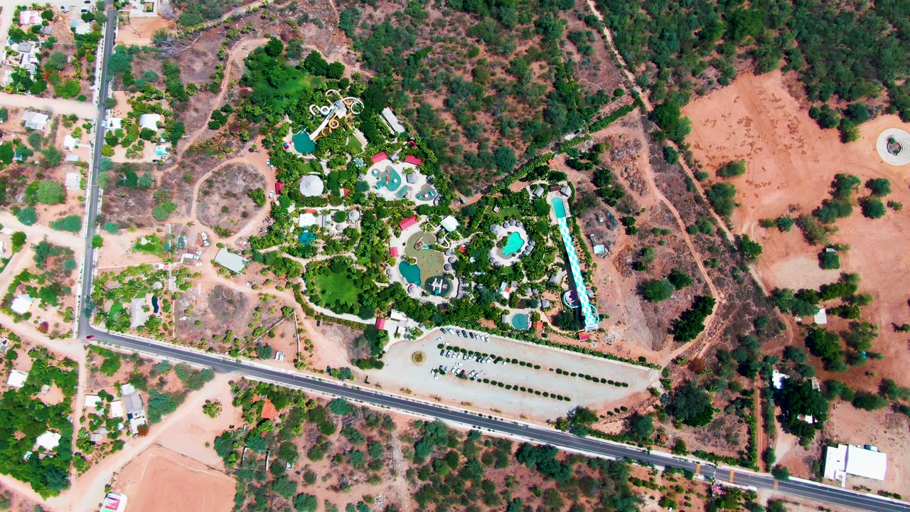 Top-down aerial frame of a resort in Caduano, Baja California Sur, with turquoise pools, dense greenery, parking rows, a paved road, and no visible people.