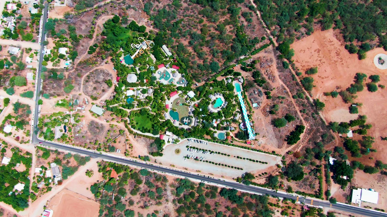 Aerial resort view with pools, greenery, parking rows, and road.