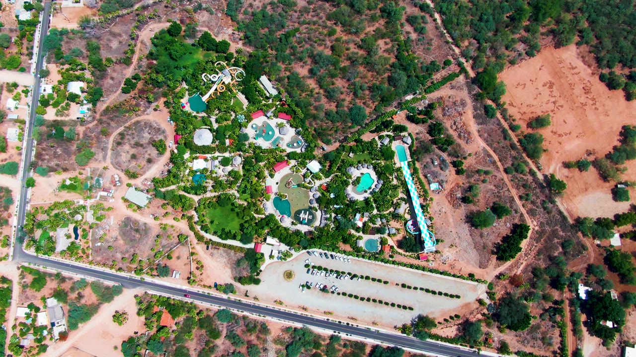 Overhead aerial frame of a Caduano resort complex with turquoise pools, a long parking area, roadside edge, surrounding trees, dry reddish terrain, and no visible people.