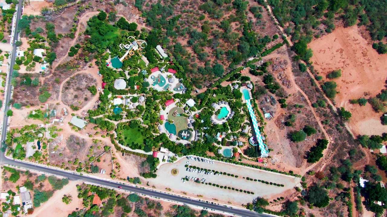 Drone view over the Caduano resort grounds showing clustered pools, pale pathways, a parking strip, a road at the lower edge, scrubby dry hillsides, and no visible people.