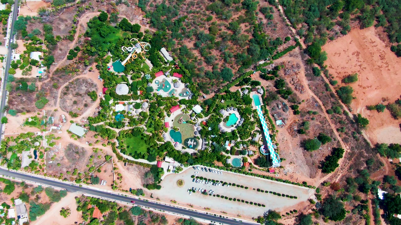 Top-down aerial frame in Caduano with the resort pools and landscaped trees bordered by a road, a light-colored parking lot, open dry terrain, and no visible people.