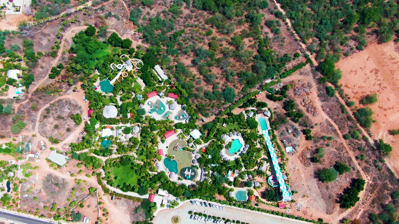 Overhead view in Caduano, Baja California Sur, showing resort pools, a long blue water slide, dense greenery, dry reddish ground, and no visible people at the opening frame.