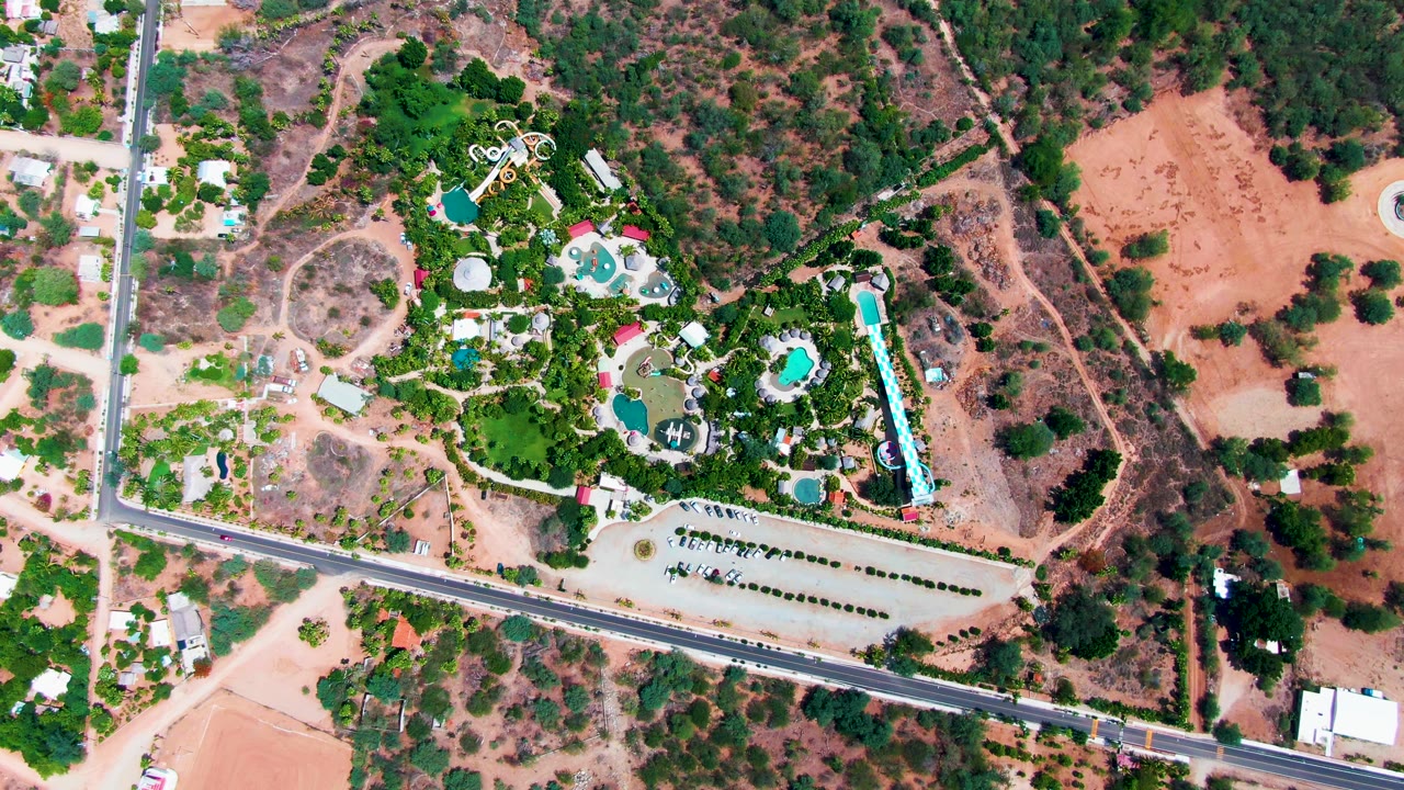 Overhead frame in Caduano, Baja California Sur showing a road below a landscaped property with pools, buildings, parked cars, trees, dry terrain, and no visible people.