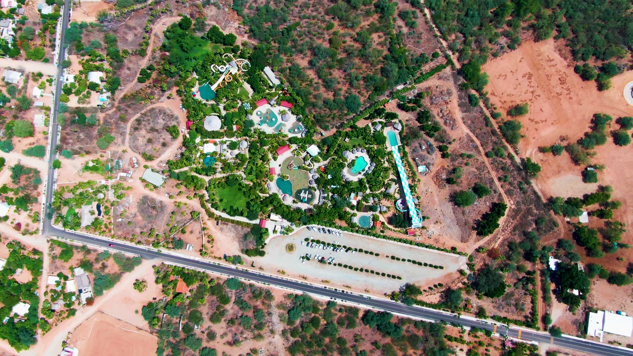 Aerial frame in Caduano, Baja California Sur with the road crossing the lower image, a small car near the right side, several pools inside a green property, dry open land, and no visible people.