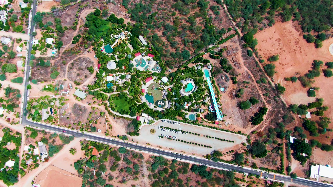 Overhead Caduano frame showing the car slightly farther left on the road, with turquoise pool shapes, dense greenery, parked cars, nearby buildings, and dry scrubland around the property, with no visible people.