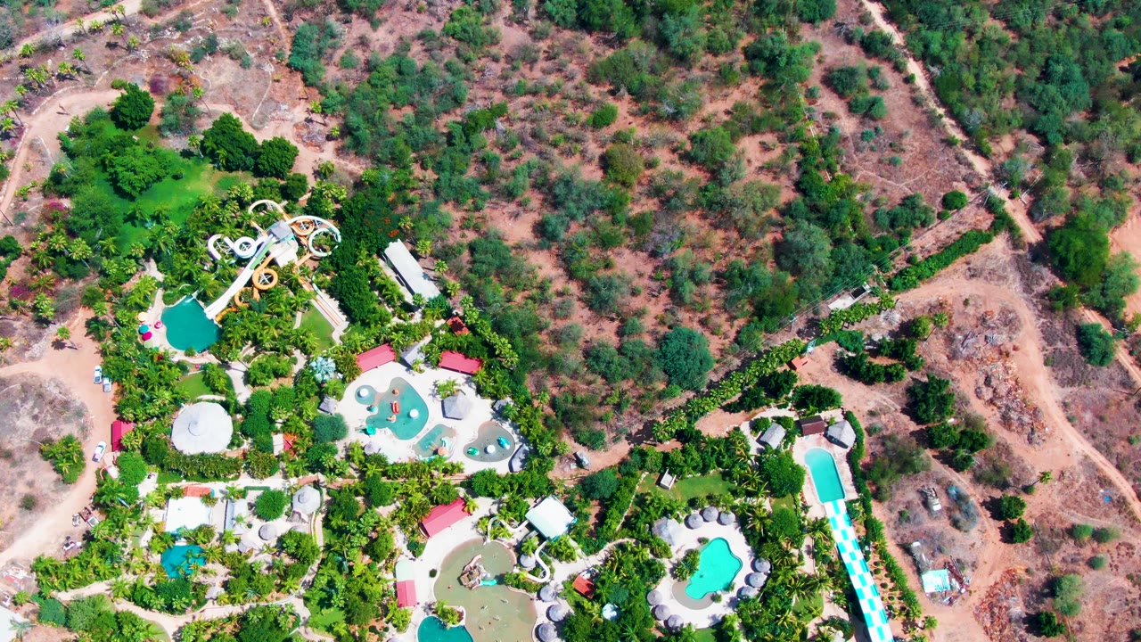 Drone view over the Caduano water park with slide towers, blue pools, planted areas, and dirt tracks crossing the dry hillside, no visible people.