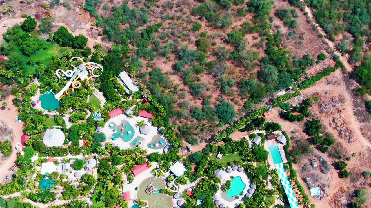Aerial frame of the Caduano water park showing bright pool shapes, white and yellow slide structures, dense green planting, and dry scrubland beyond, no visible people.