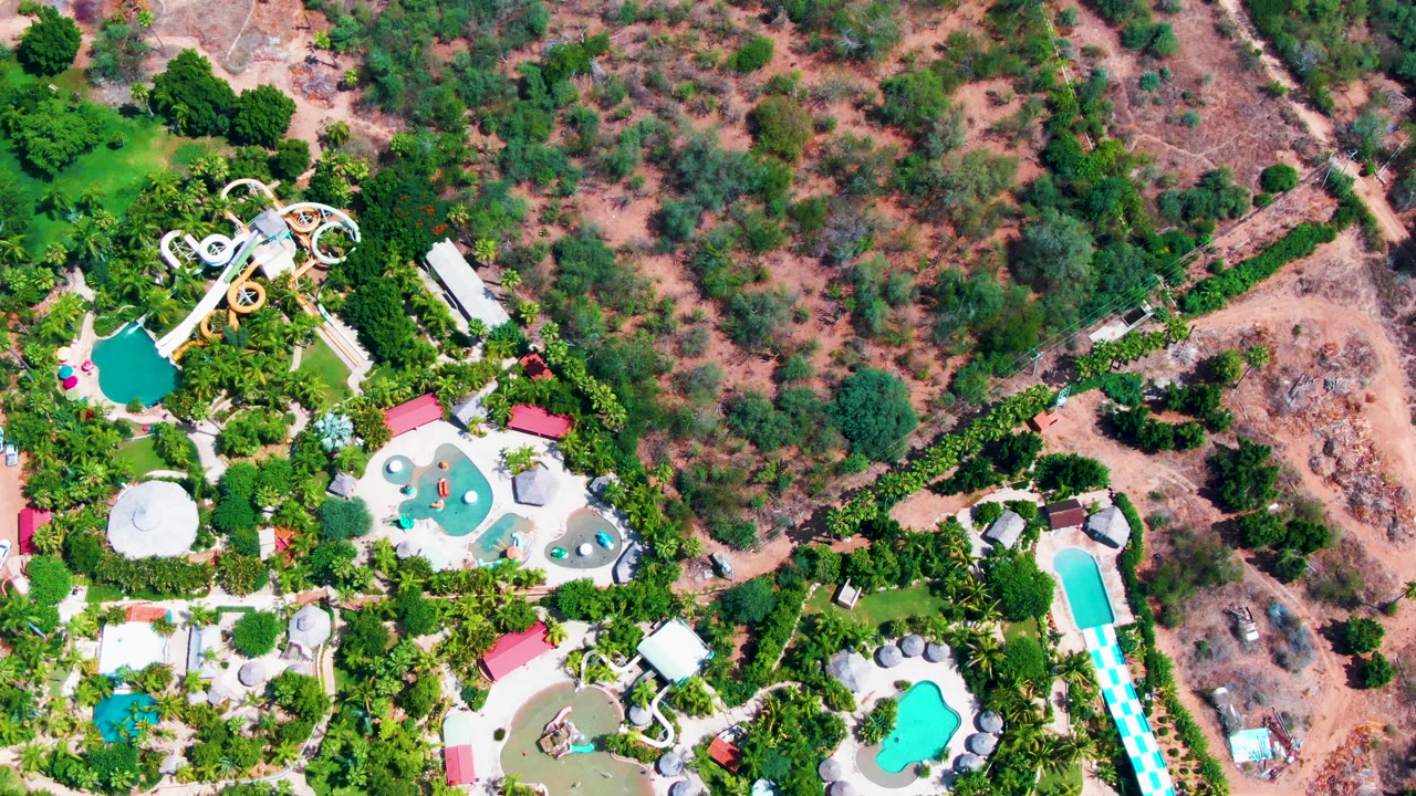Later overhead view of the Caduano site with multiple turquoise pools, palm-lined paths, slide structures, and reddish dry ground at the edge of frame, no visible people.