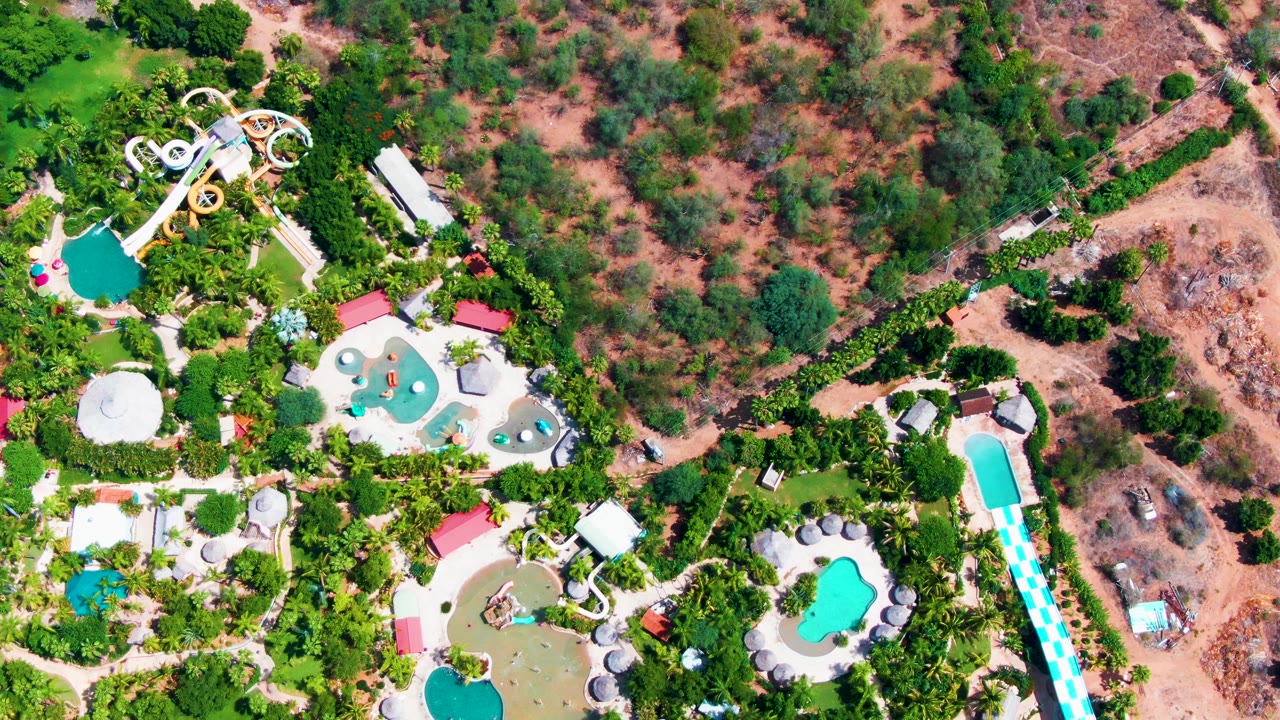High overhead view of a Caduano water park in Baja California Sur with looping slides at upper left, several pools, palms, and dry scrubland beyond.