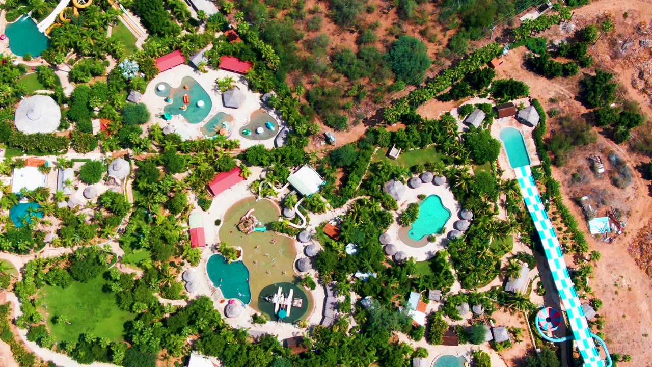Aerial view in Caduano, Baja California Sur, showing the long blue-and-white slide more centrally at right, clustered pools, white paths, heavy tree cover, and no visible people.