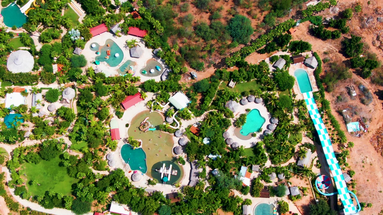 Top-down frame from Caduano, Baja California Sur, with irregular turquoise pools, a darker green pool, the checkered slide along the right edge, landscaped trees, and no visible people.