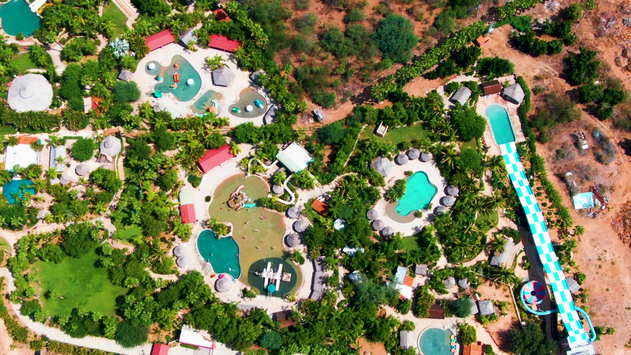 Later overhead frame in Caduano, Baja California Sur, showing the same water park from above with the long slide at right, several pools across the center, trees filling the left side, and no visible people.