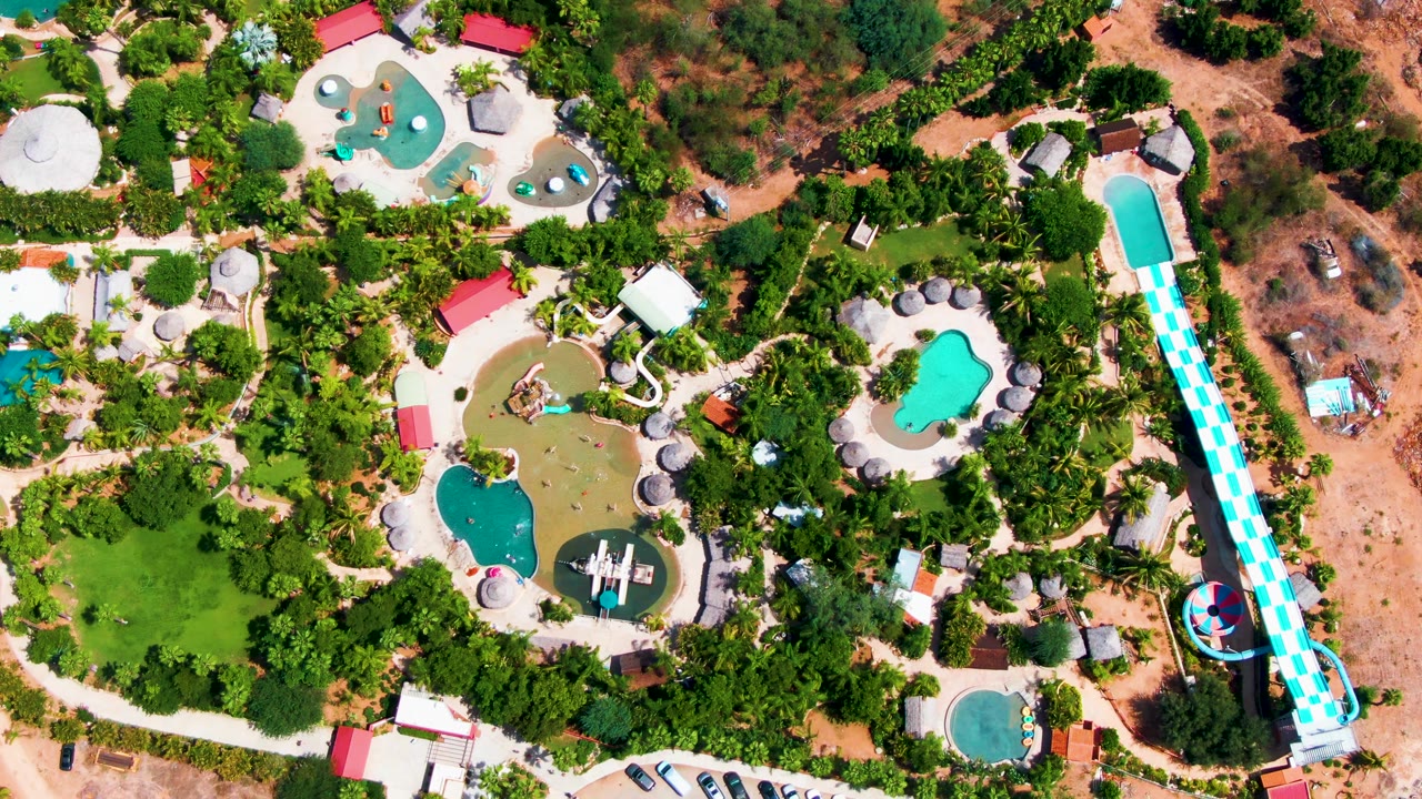 Overhead water park view with pools, trees, parking, and a long blue-and-white slide.