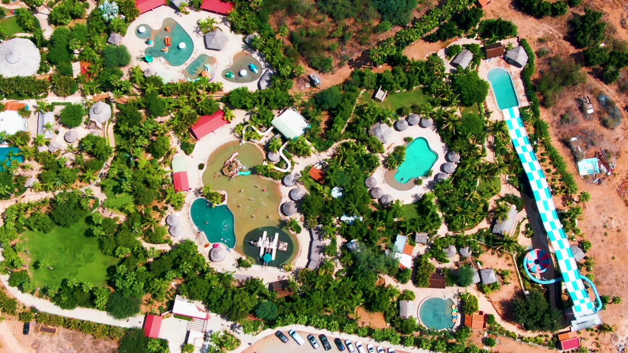 Overhead frame one second into the clip shows the Caduano water park from a similar high angle, with the long slide along the right edge, pool basins near center, parked cars at the lower edge, and no visible people.