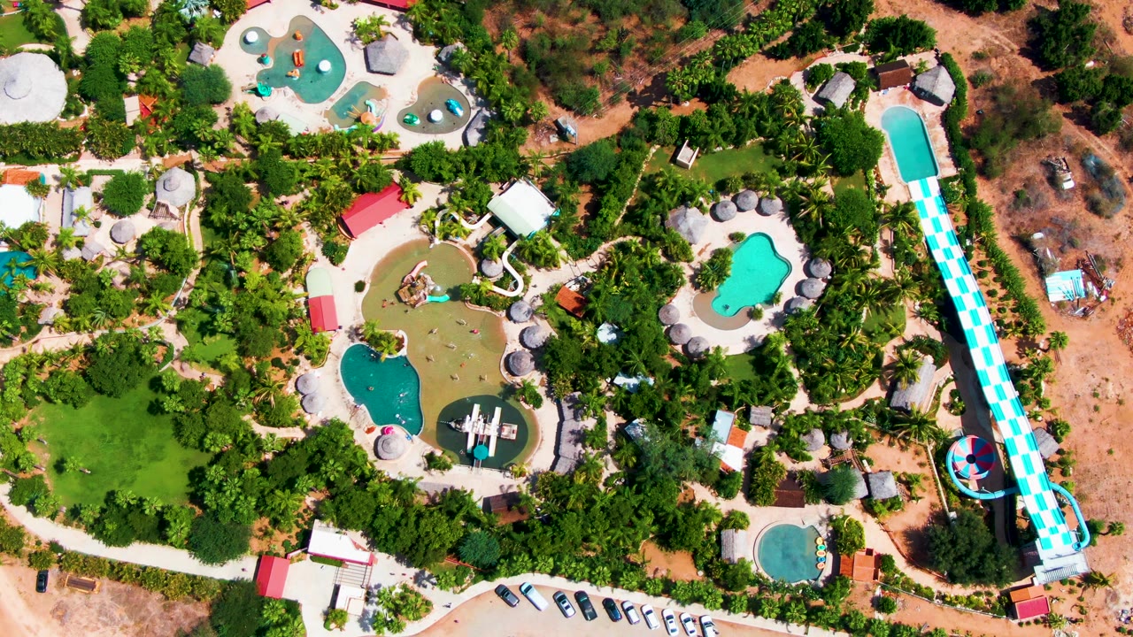 Top-down Caduano water park frame with clustered turquoise and greenish pools, small shade umbrellas, surrounding trees, parked vehicles near the bottom, and the long blue-and-white slide running vertically at right, with no visible people.