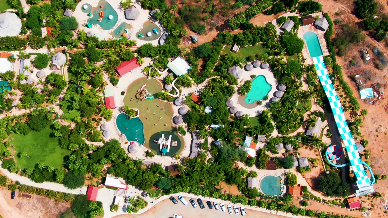 Aerial frame at 0:03 shows the long water slide near the right side, pools and paths across the center, a row of parked cars near the lower edge, dry open ground to the right, and no visible people in the Caduano park.
