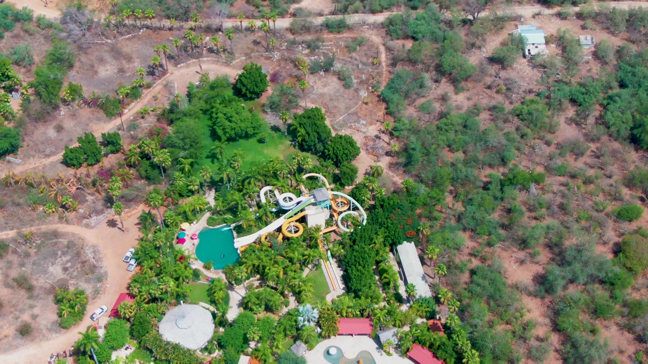 High overhead frame of a Caduano water park with turquoise pools, pale slides, green trees, dirt roads, and dry scrubland, with no visible people.