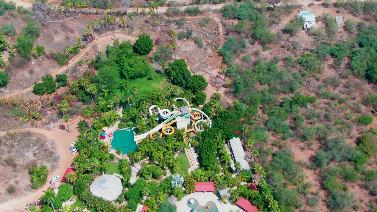 Overhead view in Caduano showing the slide complex and pool slightly farther across frame, bordered by trees, dirt tracks, scattered small structures, and no visible people.