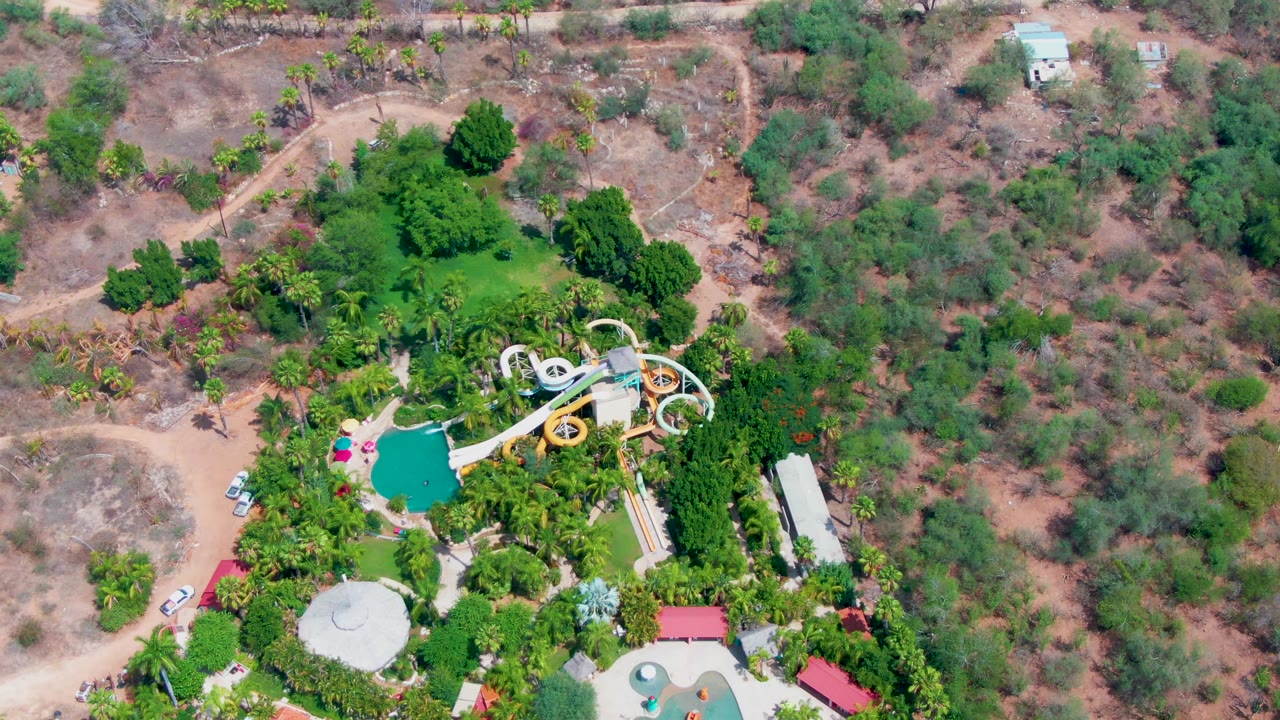 High drone view of the Caduano water park with curving slides, a bright pool, dense planted greenery, pale paths, dry surrounding terrain, and no visible people.