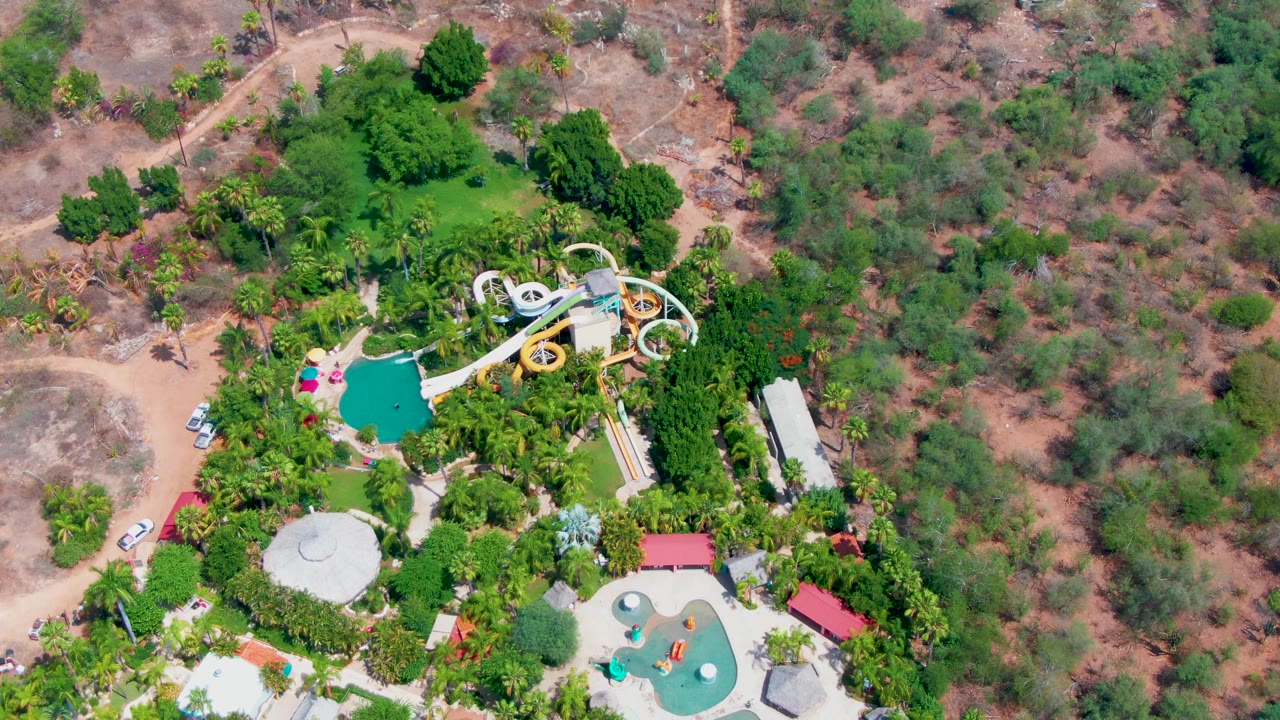 High aerial frame over a Caduano water park in Baja California Sur, showing turquoise pools, white looping slides, palms, dry scrubland, and a dirt road with small vehicles near the park edge.